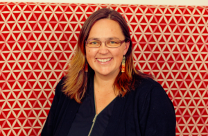 Alice Te Punga Somerville smiles in front of a red background covered in white triangles arranged into hexagons. She has brown hair that passes her shoulders, and wears purple meta-framed glasses, dangling earrings with printed green and red colours, and a black shirt with a zipper in front.