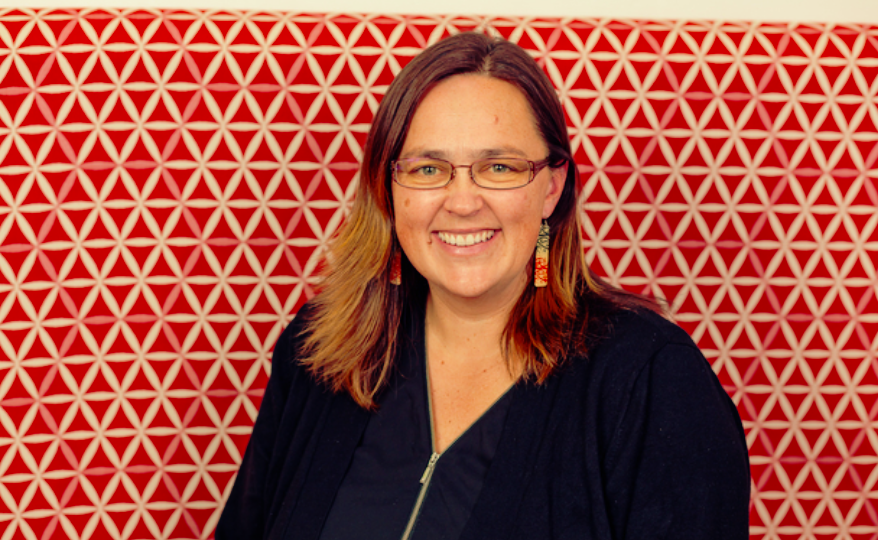 Alice Te Punga Somerville smiles in front of a red background covered in white triangles arranged into hexagons. She has brown hair that passes her shoulders, and wears purple meta-framed glasses, dangling earrings with printed green and red colours, and a black shirt with a zipper in front.