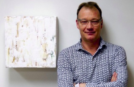 Stephen Guy-Bray posing with his arms crossed to the right of a square painting. He is wearing a dress shirt with the top button undone covered in intricate black and white fine print patterns. He is smiling.