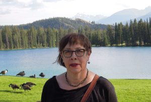 Angela Kaija posing in front of a lake, outlined with trees and an inkling of mountains in the background. The grass behind her is vividly green with a scattering of grazing Canada geese. Angela is wearing a black shirt with a a braided necklace. She wears a dangling circular earring on her left ear and dark rimmed glasses.