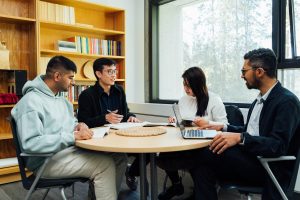 Students sit around a round table discussing books against a backdrop of shelves filled with books.